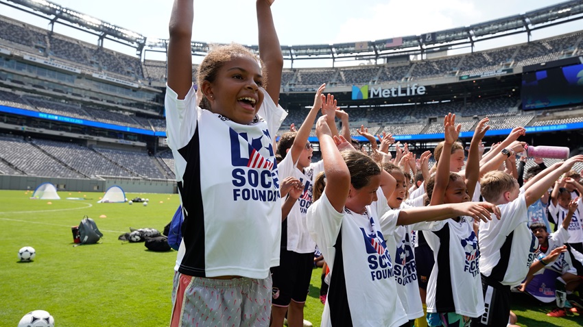 a group of kids are smiling with their hands up. they are wearing white U.S. Soccer Foundation jerseys. one child is the main focus of the image. she is smiling with her hands up in celebration during a bright, sunny day at metlife stadium on the soccer field.